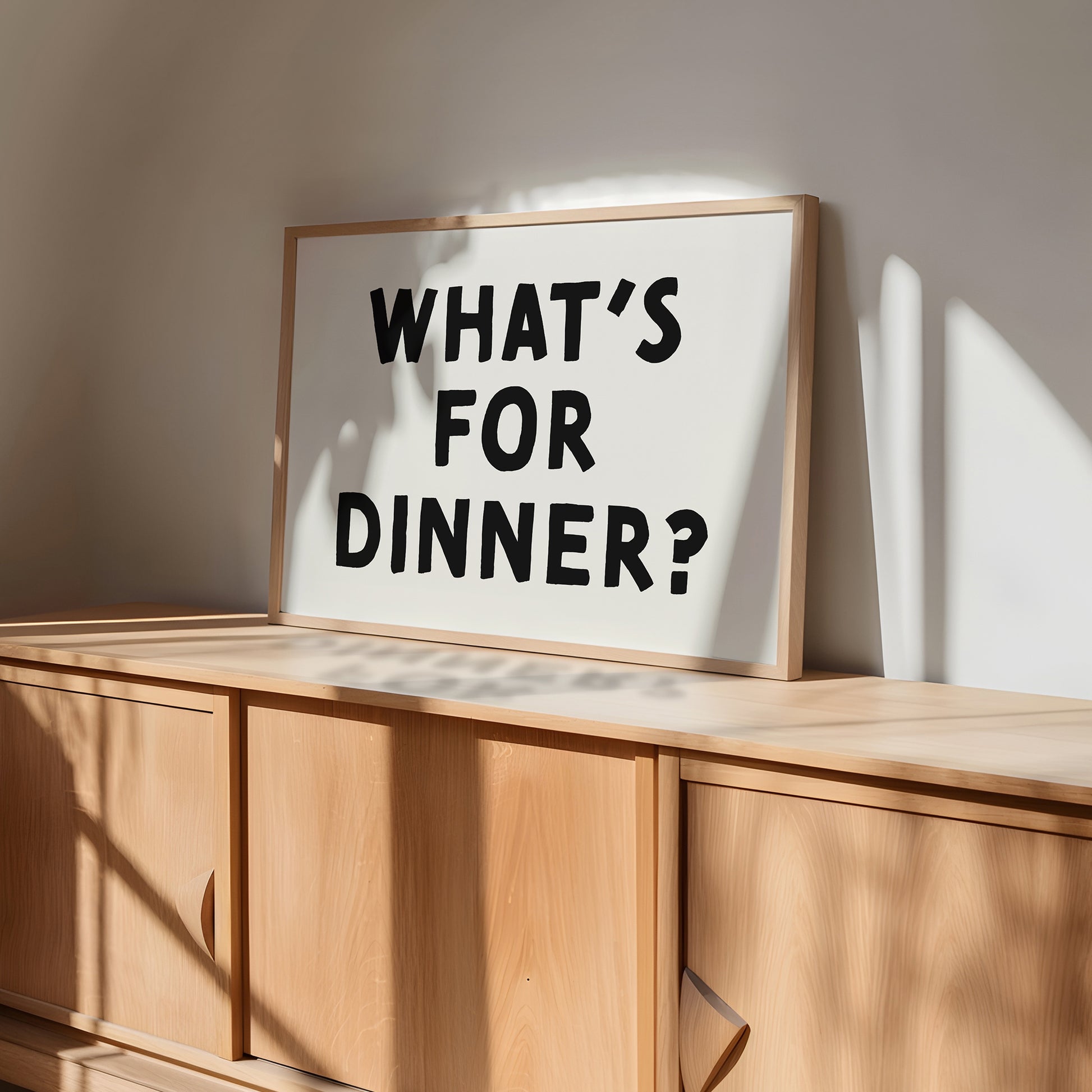 Minimalist black-and-white "WHAT'S FOR DiNNER?" poster leaning on wooden cabinet in a bright modern room.