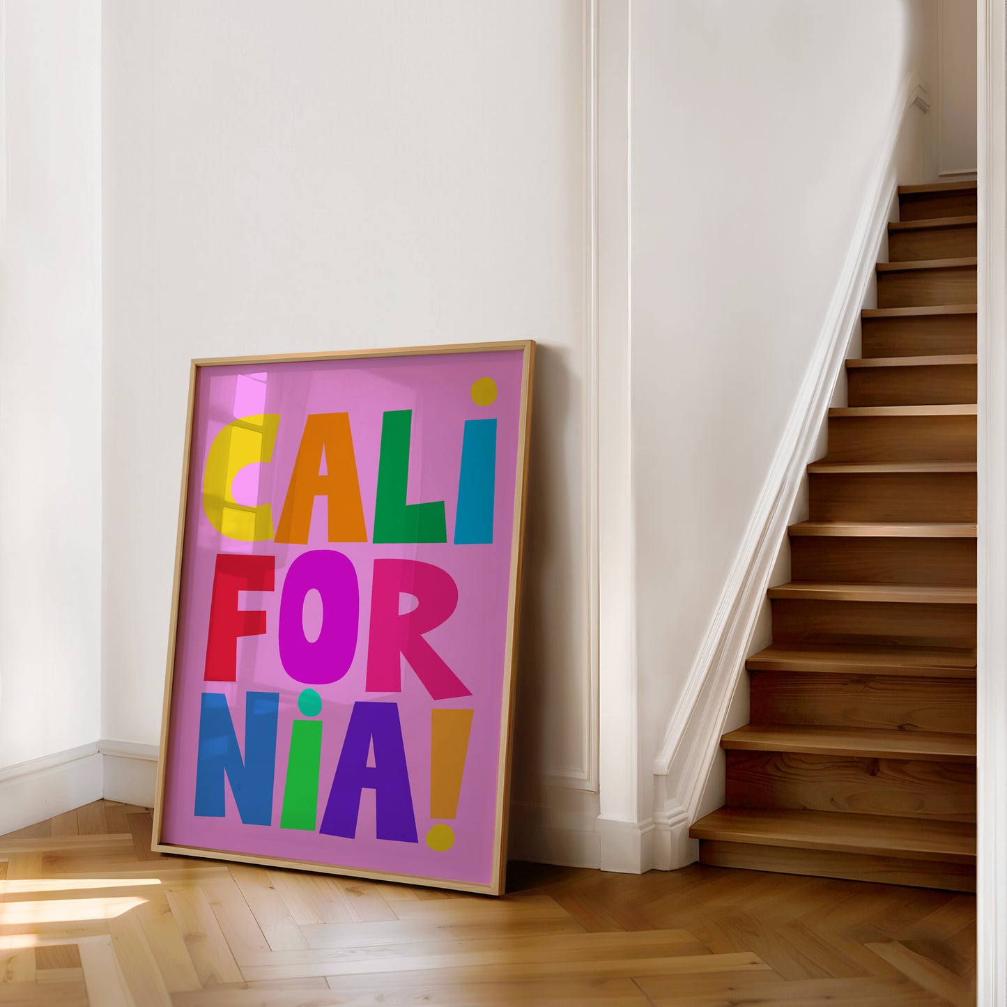 CALiFORNiA! poster with colorful block letters on a bright pink background, displayed on a wooden floor by stairs.