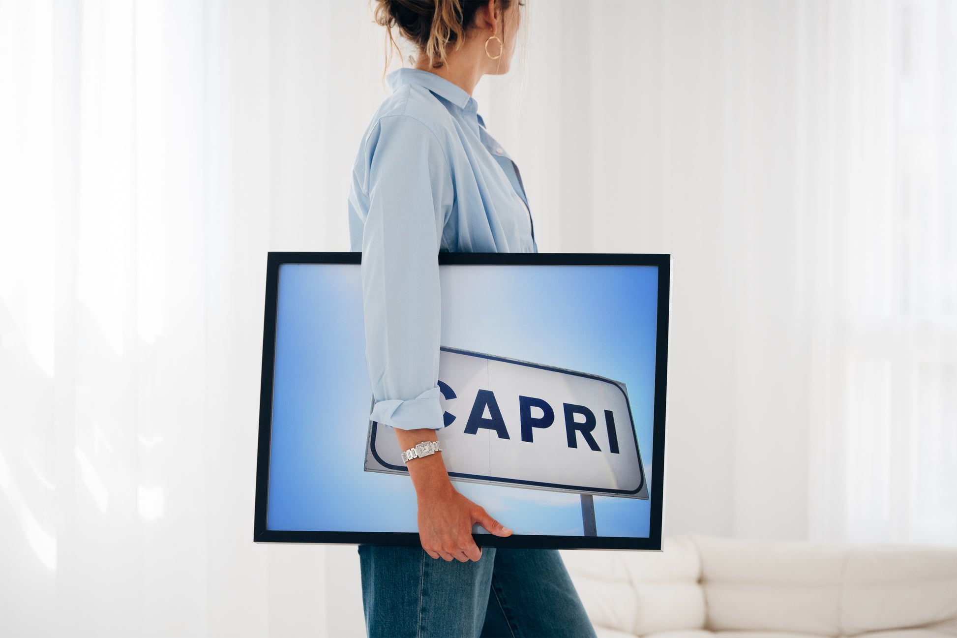 Person holding framed CAPRi sign print with bright Mediterranean sky background for home decor.