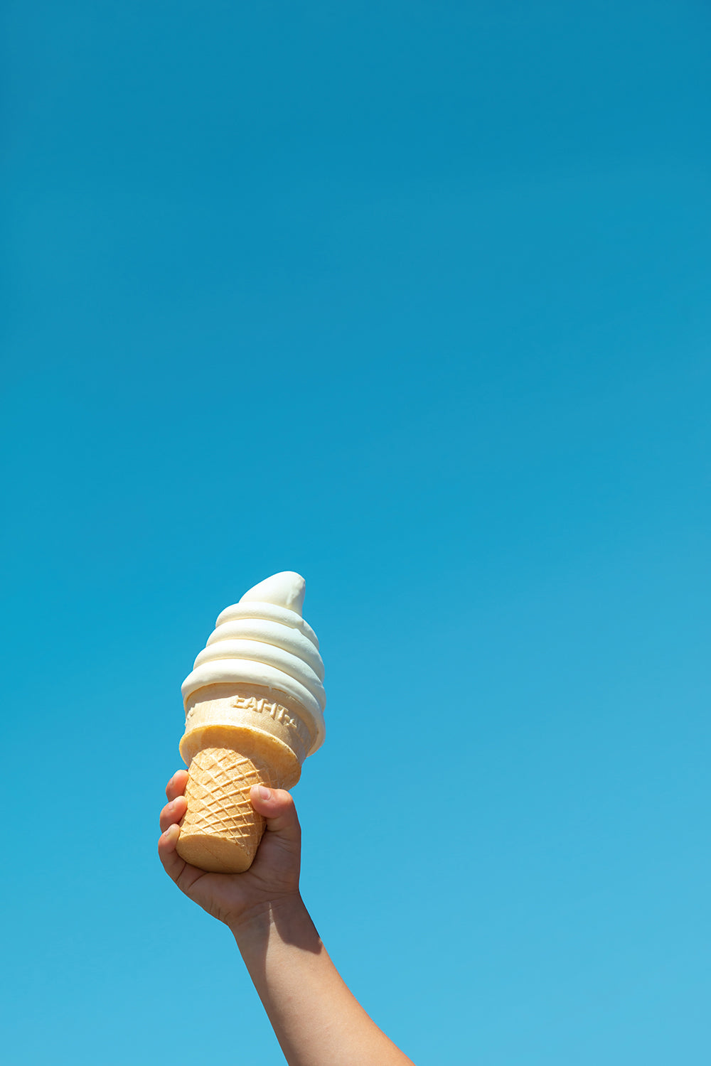 Child's hand holding soft-serve cone against clear blue sky in the product "IN FROSTiES WE TRUST" print.