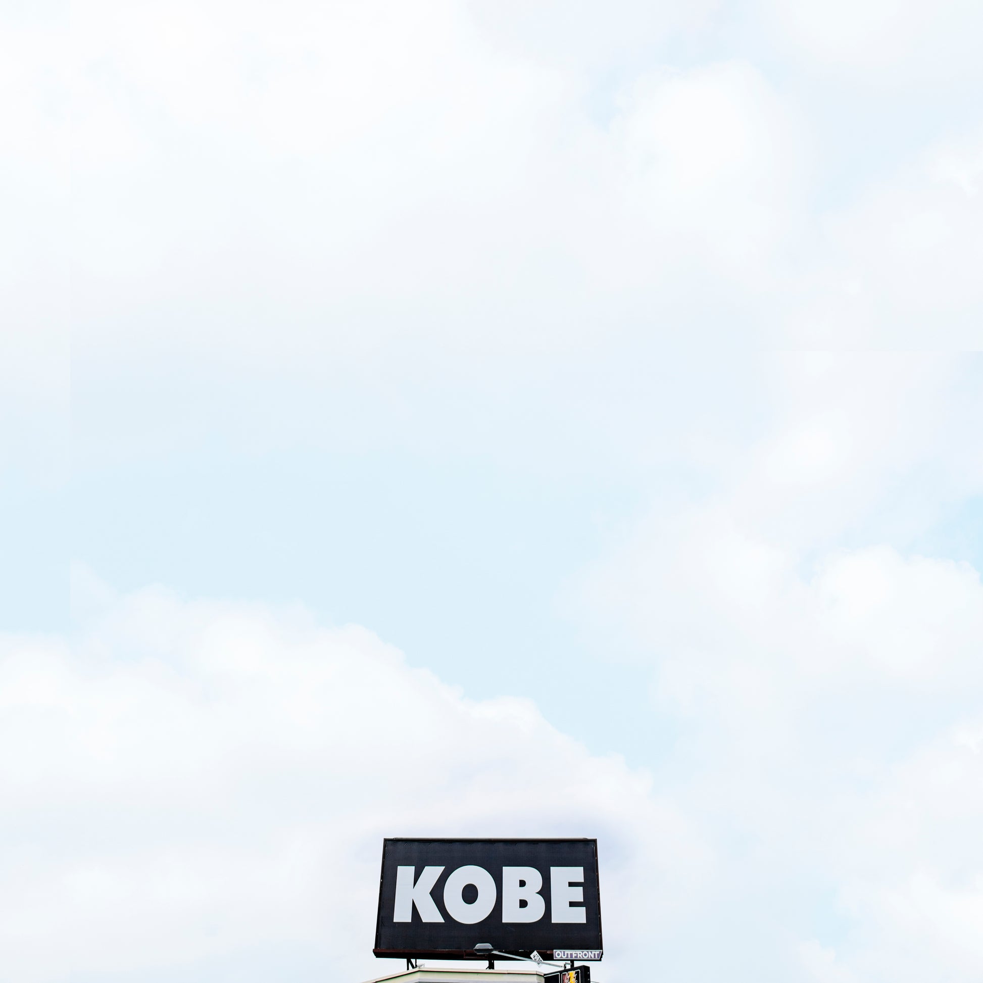 Minimalist photograph titled KOBE showing a lone urban billboard with bold white text against a cloudy sky.