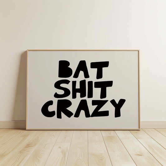 Framed black and white poster with chunky hand-lettered typography reading BAT SHiT CRAZY displayed on wooden floor.