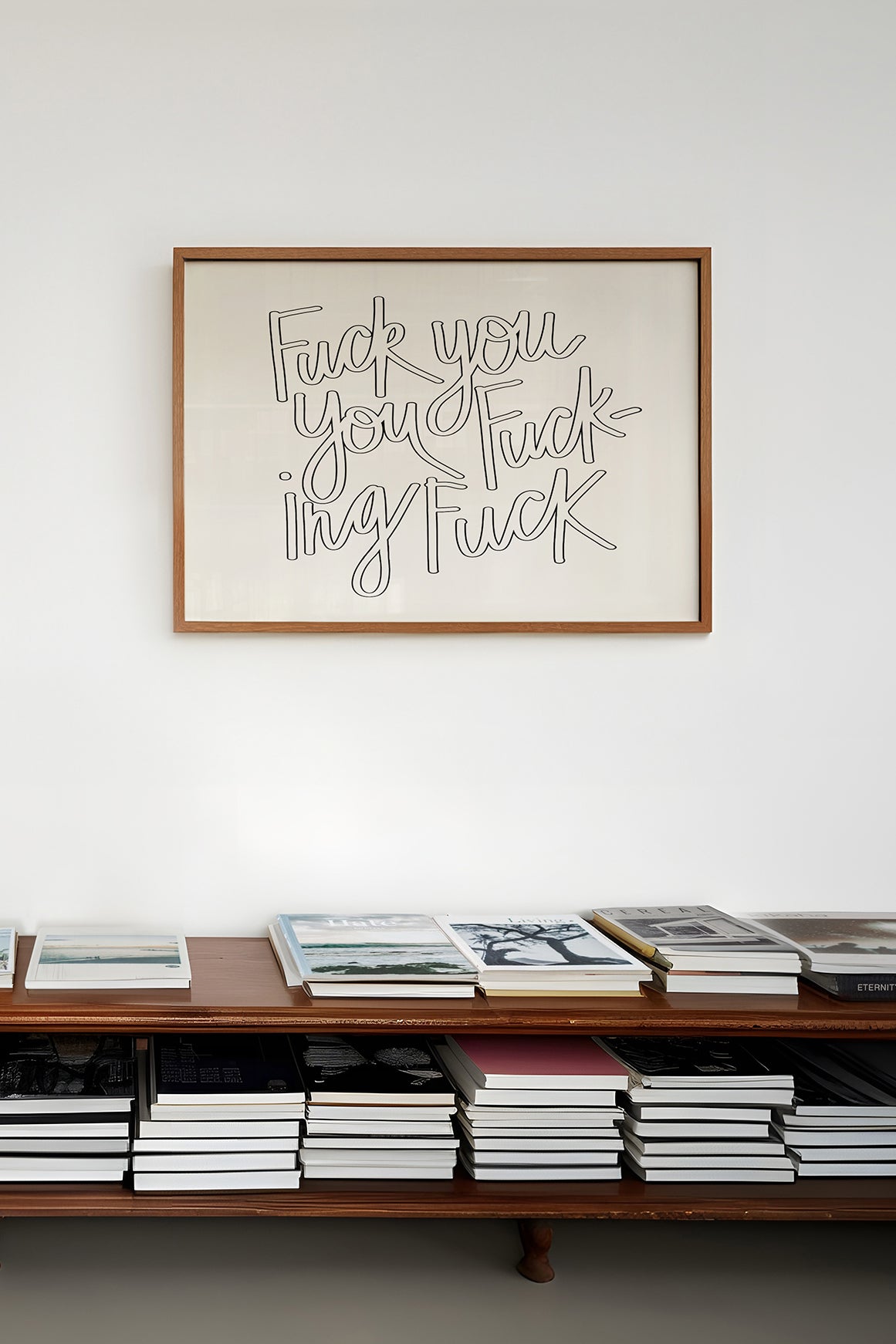 Framed black-and-white hand-lettered poster titled "FUCK YOU YOU FUCKING FUCK" displayed above book-filled wooden shelf.