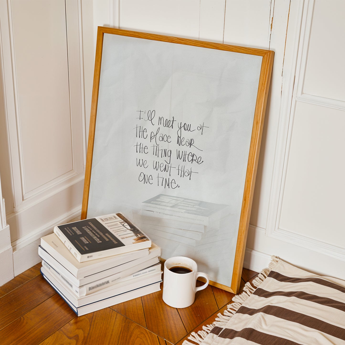 Framed I’LL MEET YOU AT THE PLACE ... poster with handwritten text, displayed by stacked books and coffee mug on wood floor.