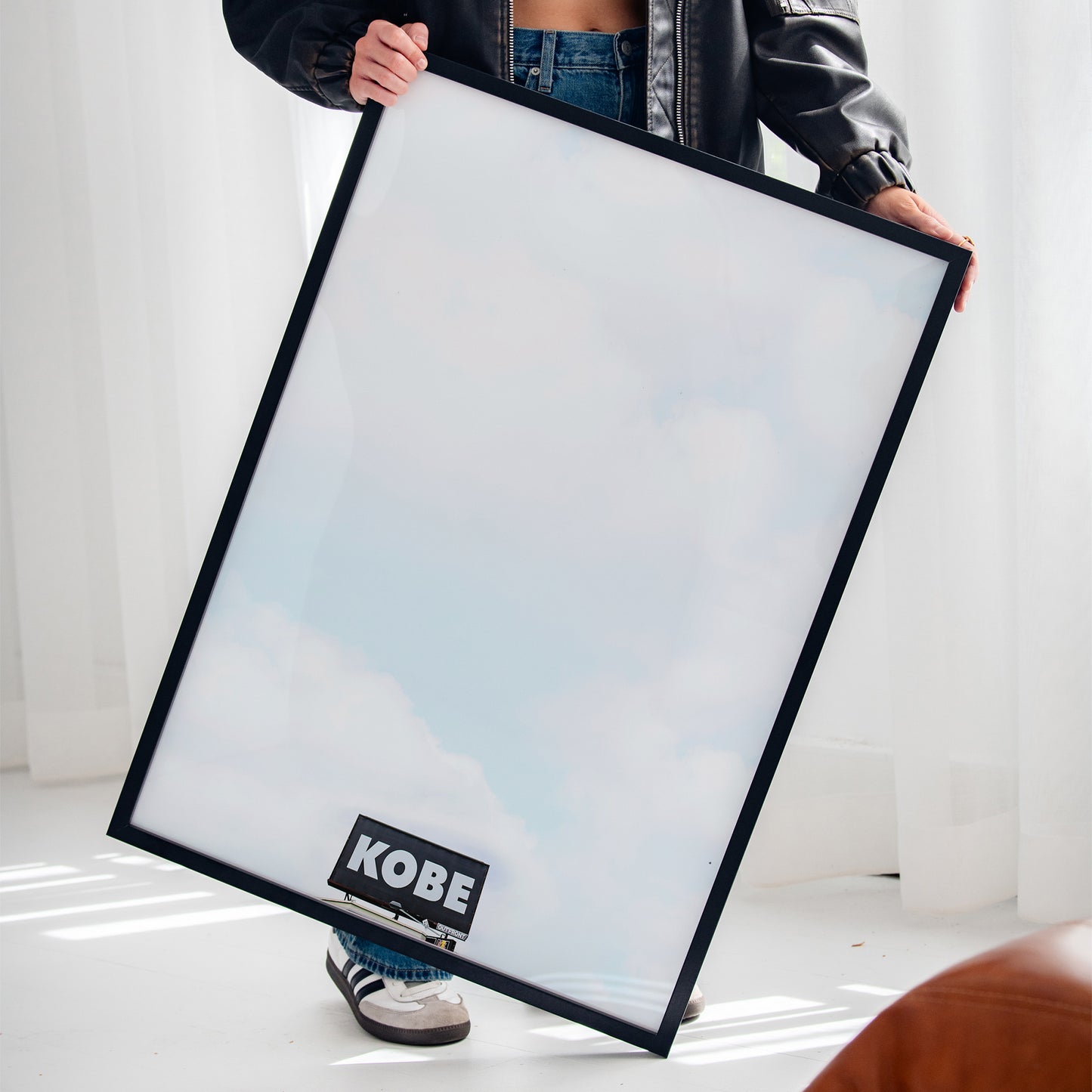 Person holding framed KOBE minimalist photograph with urban billboard and cloudy sky, vertical art print.