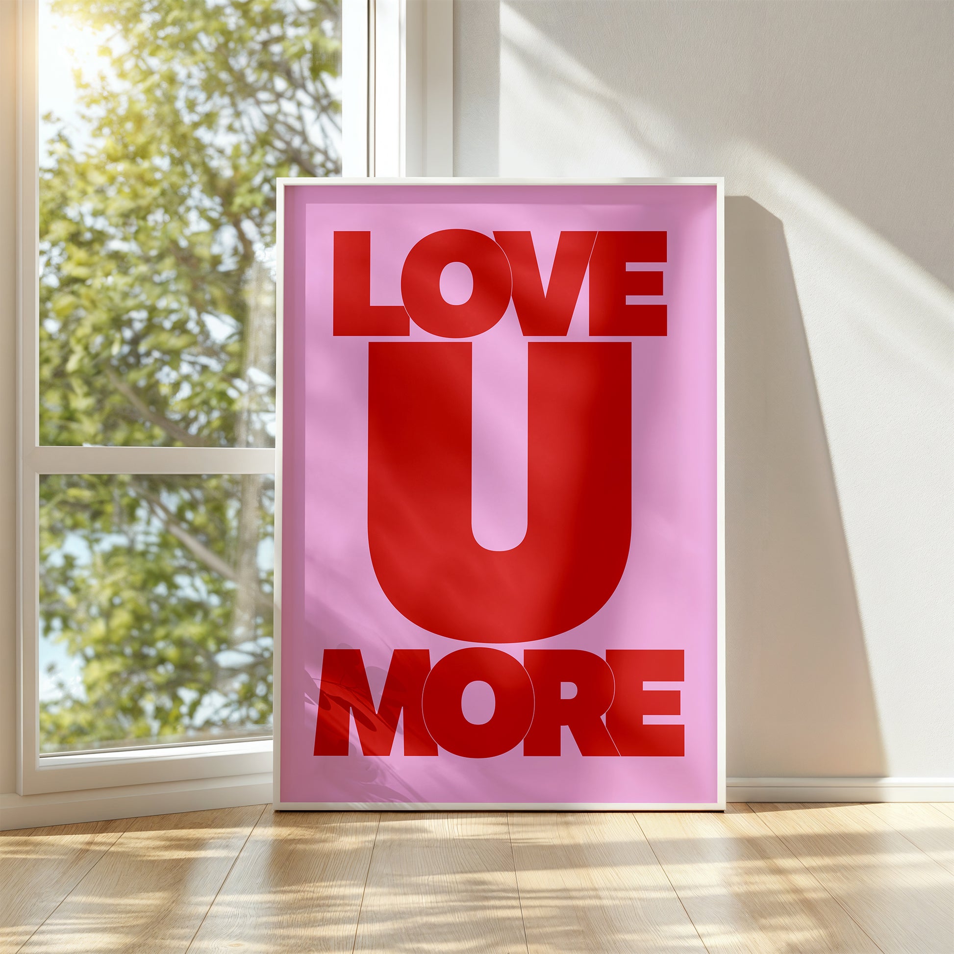 LOVE U MORE poster with bold red typography on vibrant pink background displayed in sunlit room.