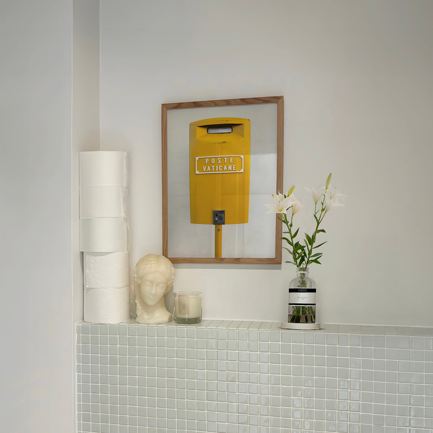 Framed "POSTE VATiCANE" bright yellow Vatican mailbox art print on wall above tiled shelf with flowers and decor.