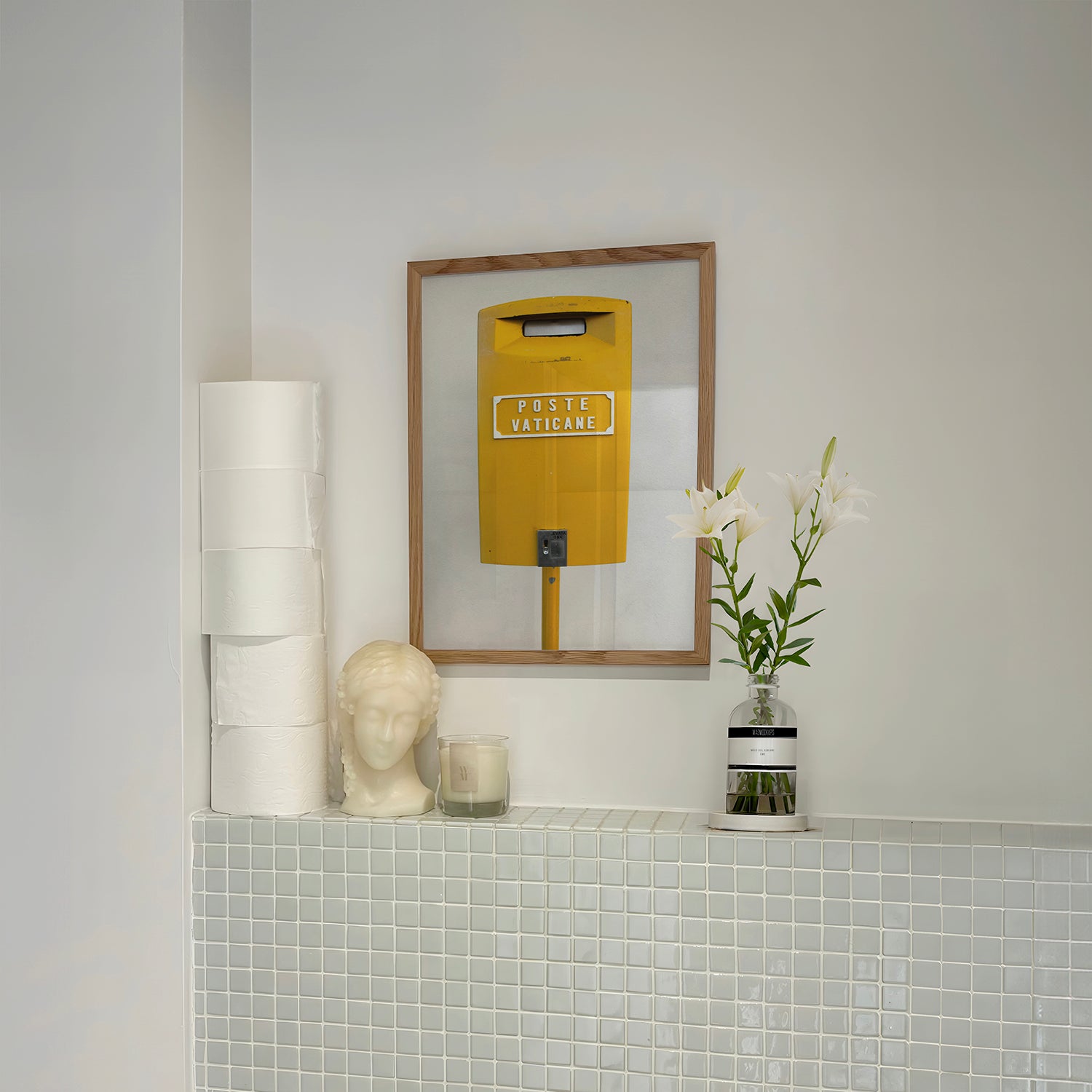 Framed "POSTE VATiCANE" bright yellow Vatican mailbox art print on wall above tiled shelf with flowers and decor.