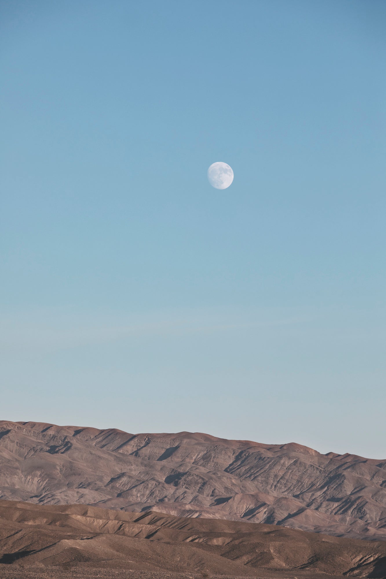RiSiNG features the moon rising above rugged desert mountains under a soft blue sky, a peaceful minimalist scene.