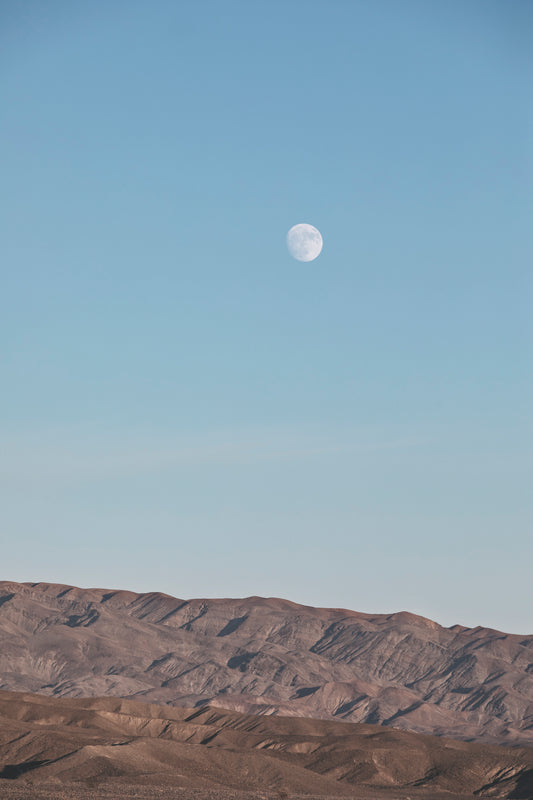 RiSiNG features the moon rising above rugged desert mountains under a soft blue sky, a peaceful minimalist scene.