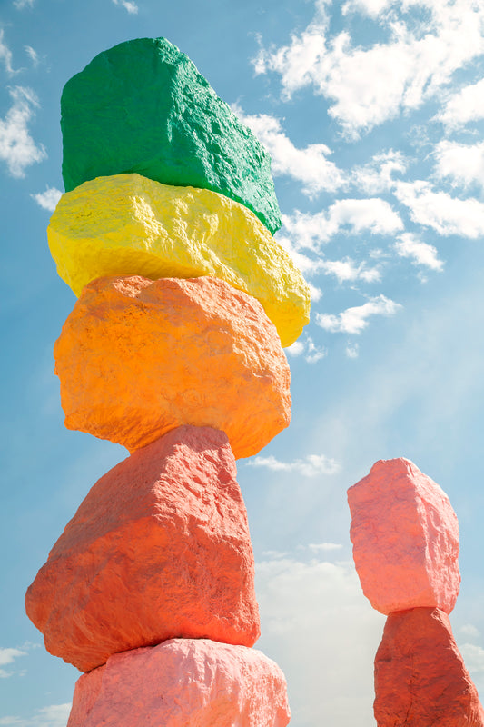 ROCK ON photograph of stacked neon-painted boulders under a bright blue sky with scattered clouds.