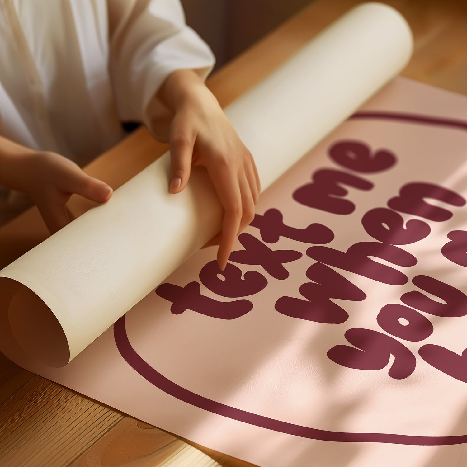 Hands unrolling a "TEXT ME WHEN YOU GET HOME" poster with burgundy typography on a blush background.