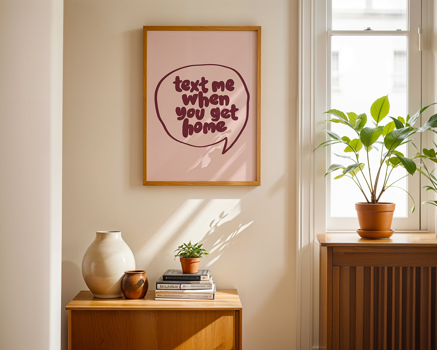 Framed "TEXT ME WHEN YOU GET HOME" poster in burgundy script on blush, styled in a cozy sunlit room with plants.