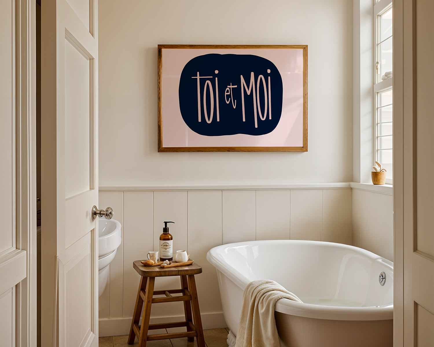 Framed "TOi et MOi" poster with blush script on navy background hanging in a bright minimalist bathroom.