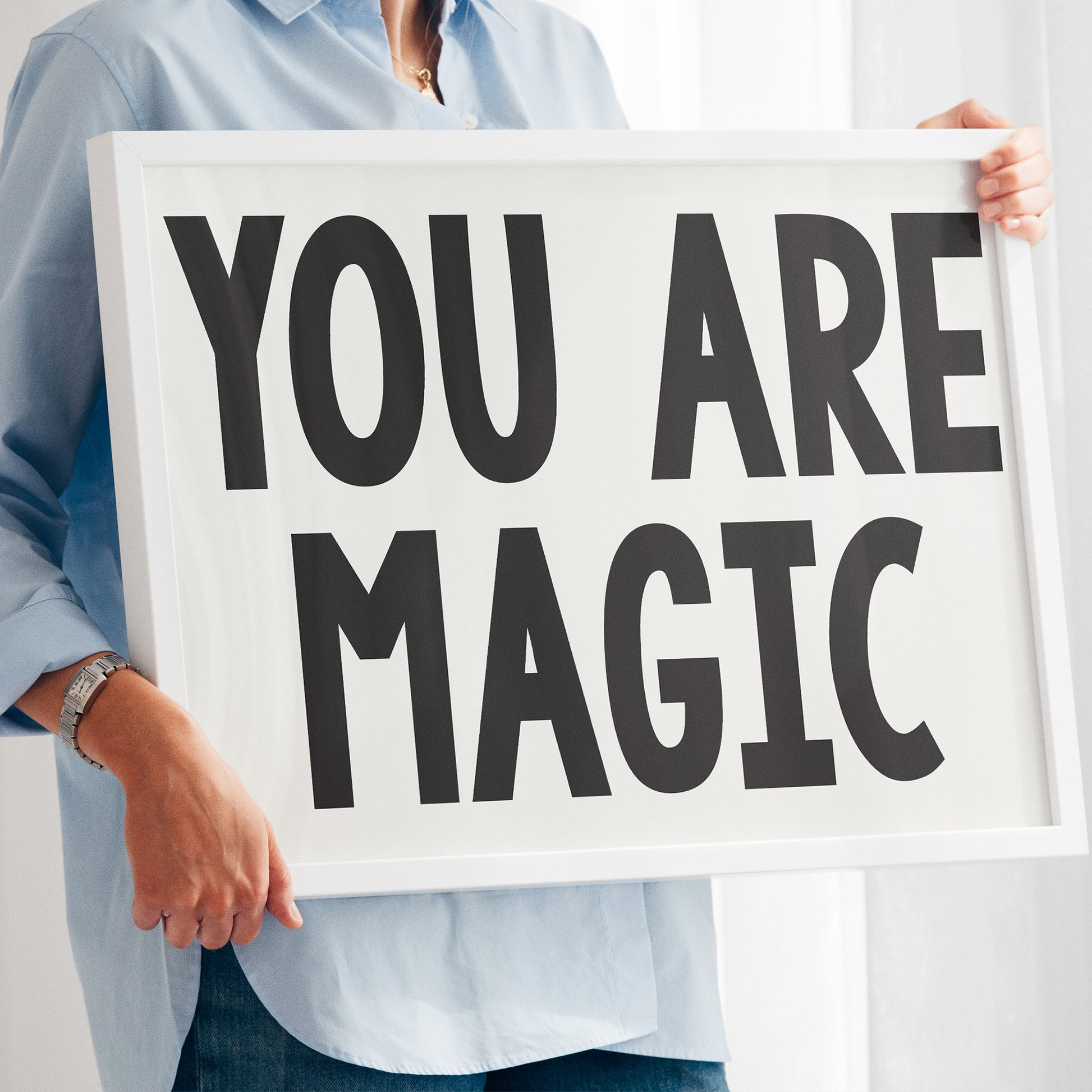 Person holding a framed typographic poster with bold text reading YOU ARE MAGiC, minimalist and stylish design.